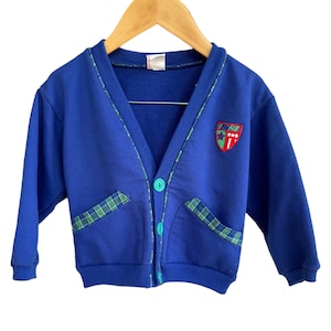 May include: A blue cardigan with a green and blue plaid trim and a red and white embroidered patch. The cardigan has a button closure and two pockets.