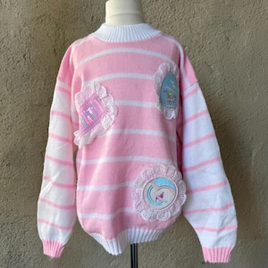 Peut inclure: Un pull rayé rose et blanc avec des écussons décoratifs. Le pull présente un col, des poignets et un ourlet côtelés blancs. Des écussons avec des bordures en dentelle et des motifs brodés sont cousus sur le devant. Le pull est présenté sur un mannequin.