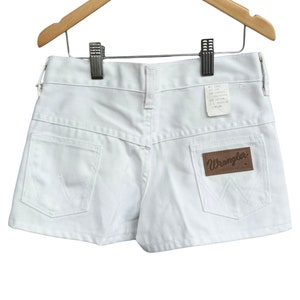 Peut inclure: Short en jean blanc avec une étiquette Wrangler en cuir marron sur la poche arrière. Le short a une taille haute et une braguette à bouton.