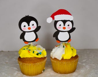 May include: Two cupcakes decorated with yellow and white frosting. Each cupcake has a penguin topper. One penguin is wearing a red Santa hat.