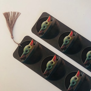 Op de afbeelding: Bladwijzers met een groen en bruin personage met grote ogen en puntige oren, tegen een donkere achtergrond. Elke bladwijzer heeft een bruine kwast bevestigd. Het personage zit in een donkere pod. De bladwijzers liggen op een wit oppervlak.