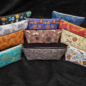 May include: A collection of 12 zippered pouches with various Harry Potter themed designs. The pouches are made of fabric and feature different colours and patterns, including a blue pouch with glasses, a red pouch with a golden snitch, and a yellow pouch with a Hogwarts crest.