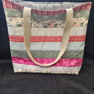 May include: A large, striped tote bag with a patchwork design. The bag is made of various fabrics in shades of green, red, pink, and blue. The bag has two long, beige straps.