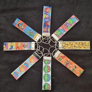 May include: Eight colourful keychains featuring cartoon characters, including the Teenage Mutant Ninja Turtles, Care Bears, and the Simpsons. The keychains are attached to silver key rings.