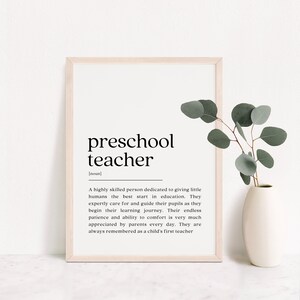 Puede incluir: Una impresión de arte imprimible en blanco y negro con la definición de un maestro de preescolar. La impresión está enmarcada en un marco de madera claro y se encuentra sobre una superficie blanca con una planta verde en un jarrón blanco.
