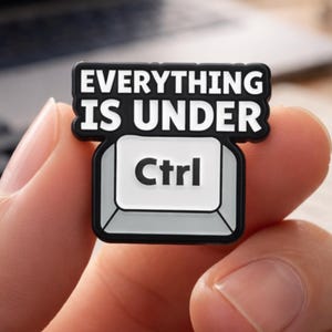 May include: A black and white charm in the shape of a computer key, with the words "EVERYTHING IS UNDER Ctrl". The charm is held in a hand, with a laptop and other items on a desk in the background.