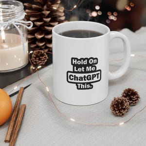 Puede incluir: Una taza de café de cerámica blanca con el texto "Hold On Let Me ChatGPT This." en negro. La taza contiene una bebida oscura y está colocada sobre una superficie gris claro, junto a piñas y una vela encendida.