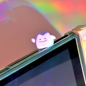 May include: A light purple Ditto-shaped accessory sits atop a black electronic device. The accessory has a smiling face and small arms. The background features a pink hue with rainbow light reflections.