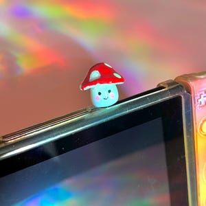 May include: A small, whimsical mushroom figurine with a red cap and white spots, perched on top of a black electronic device. The mushroom has a light blue base with a smiling face. The background has a soft pink hue with rainbow light reflections.