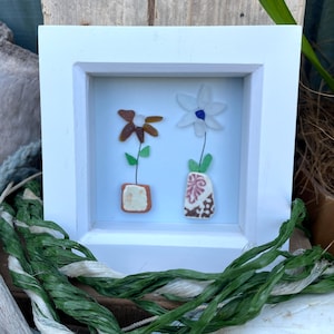 May include: A white framed picture featuring two sea glass flowers in small pots. The flowers are brown and white with green leaves. The pots are decorated with brown and white patterns.