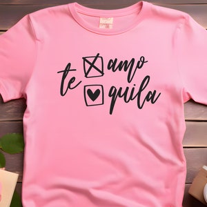 Op de afbeelding: Roze T-shirt met zwarte tekst die "te amo" zegt met een kruissymbool en "quila" met een hartje.