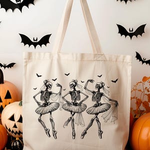 May include: A white canvas tote bag with a black and white illustration of three skeletons dressed as ballerinas. The skeletons are wearing tutus and have bows in their hair. There are small black bats scattered around the skeletons.