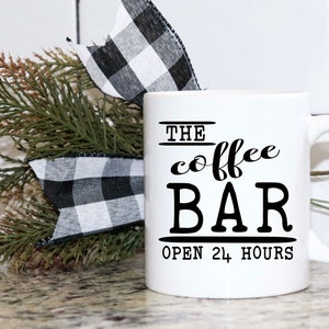 May include: White ceramic coffee mug with black text that reads "The coffee bar open 24 hours".