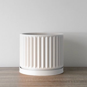May include: A white, cylindrical planter with vertical ribbed detailing. The planter sits on a matching round saucer. The design is modern and minimalist, suitable for indoor or outdoor use. The planter is made of ceramic.