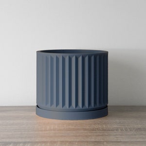 May include: A dark blue ceramic planter with a ribbed design and a matching saucer. The planter is sitting on a wooden surface.