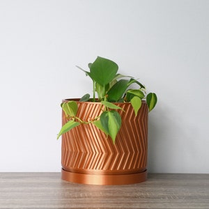 Puede incluir: Una maceta de color cobre con un patrón geométrico contiene una planta verde con enredaderas colgantes. La maceta tiene una base a juego de color cobre.
