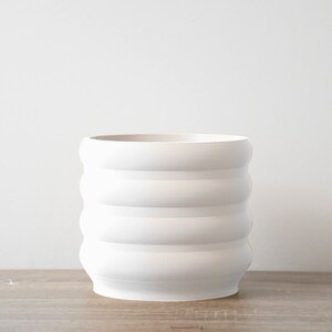 May include: White ceramic planter with a ridged design. The planter has four horizontal ridges.
