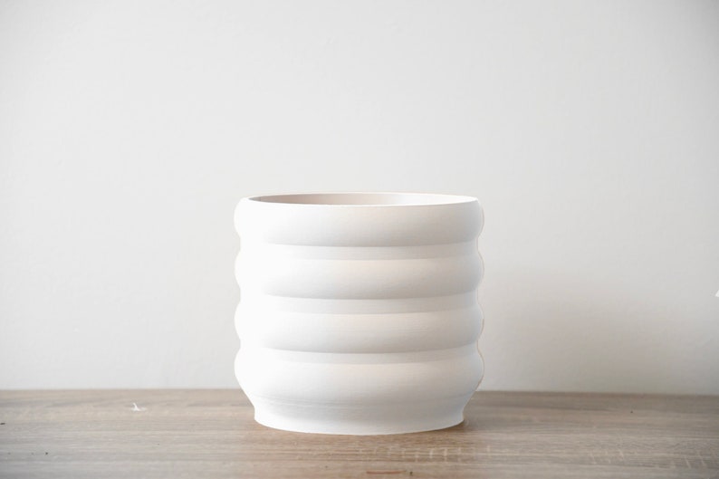May include: White ceramic planter with a ridged design. The planter has four horizontal ridges.