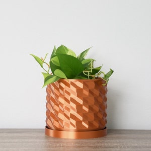 May include: A copper-colored geometric planter with a green plant inside. The planter has a textured, diamond-shaped pattern.