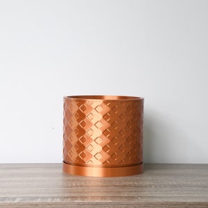 May include: A copper-colored planter with a geometric diamond pattern. The planter has a matching saucer.