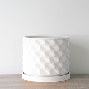 May include: White ceramic planter with a geometric pattern and a matching saucer. The planter is designed to hold a small plant or flower.