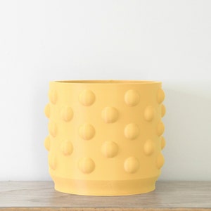 May include: A yellow ceramic planter with a textured, dotted surface. The planter is round and has a smooth, rounded bottom.