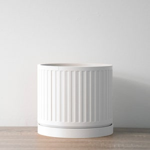 May include: White ceramic planter with a ribbed design and a matching saucer. The planter is empty and sitting on a wooden surface.