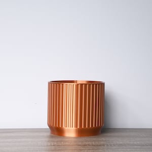Peut inclure: Un pot de fleurs de couleur cuivre avec un design côtelé. Le pot est posé sur une surface en bois.