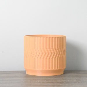Op de afbeelding: Een terracottakleurige plantenpot met een geometrisch patroon. De pot is cilindrisch en heeft een glad, licht getextureerd oppervlak.