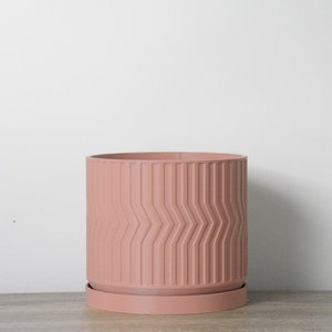Puede incluir: Una maceta de color rosa polvoriento con un patrón geométrico y un platillo a juego. La maceta está hecha de un material liso y duradero.