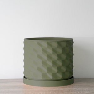 May include: A green ceramic planter with a geometric pattern. The planter has a wide, round base and a slightly narrower top. The planter is sitting on a wooden surface.