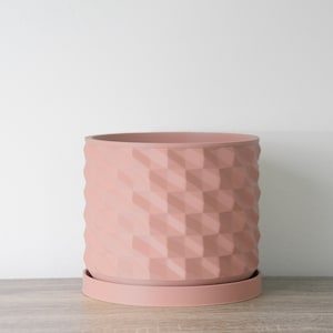 May include: A blush pink planter with a geometric pattern and a matching saucer. The planter is made of a textured material and has a modern design.