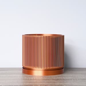 May include: A copper-colored cylindrical planter with vertical ribbed detailing. The planter sits on a matching saucer, and is placed on a wooden surface against a white background. This decorative planter is suitable for indoor or outdoor use.