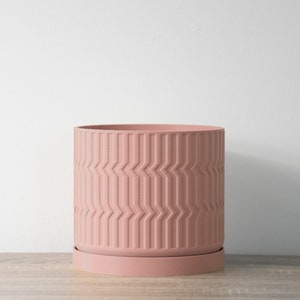 Może przedstawiać: Różowa doniczka z geometrycznym wzorem i pasującą podstawką. Doniczka wykonana jest z gładkiego, trwałego materiału.