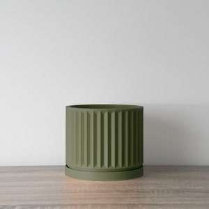 May include: A green ceramic planter with a ribbed design and a matching saucer. The planter is sitting on a wooden surface.