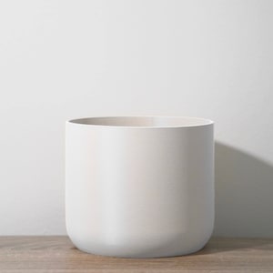 以下が含まれることがあります： 丸みを帯びた底を持つシンプルな白いセラミックプランター。プランターは木製の表面の上に置かれています