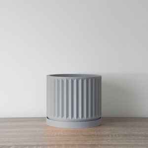 May include: A gray ceramic planter with a ribbed design and a matching saucer. The planter is sitting on a wooden surface.