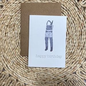 May include: A white card with a drawing of a pair of waders and the text "happy birthday". The card is on a brown envelope and a woven straw mat.