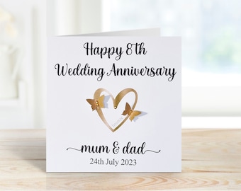 May include: A white card with gold lettering that reads "Happy 8th Wedding Anniversary" with a gold heart and two gold butterflies. The text "mum & dad" is written in cursive below the heart and the date "24th July 2023" is written below that.