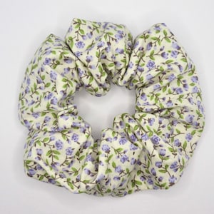 Puede incluir: Un scrunchie blanco con un patrón floral con pequeñas flores moradas y hojas verdes.