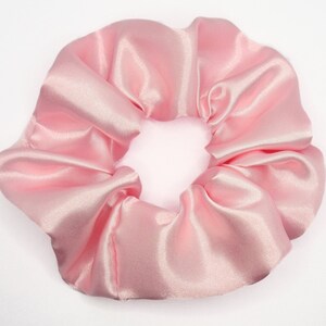 May include: A light pink hair scrunchie with a smooth, shiny, satin-like finish. The fabric is gathered into soft, voluminous folds around a circular elastic band, creating a rounded, ruffled appearance with a consistent pastel pink color.