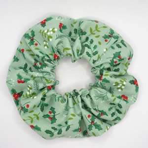 Può includere: Un elastico per capelli verde chiaro con un motivo ripetuto di foglie di agrifoglio e bacche rosse. Il tessuto è raccolto attorno a un'apertura centrale, creando una forma circolare e gonfia. Accessorio per capelli.