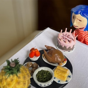 Puede incluir: Una muñeca en miniatura con cabello azul y una camisa a rayas está sentada cerca de una mesa con un festín en miniatura. La mesa incluye un pavo asado, pastel con el texto "welcome home", maíz, guisantes, patatas y una piña.
