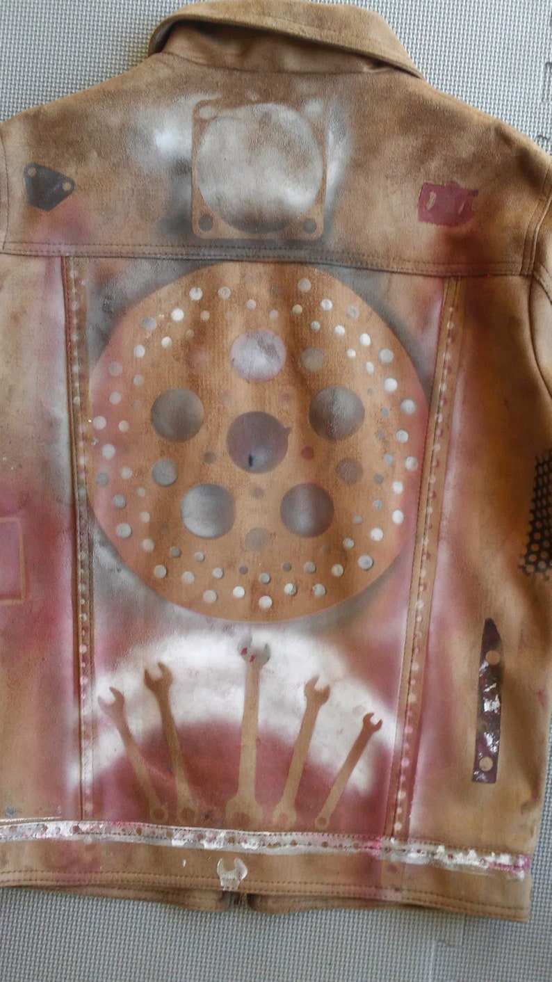 Puede incluir: Una chaqueta de ante marr&oacute;n con un dise&ntilde;o pintado con aerosol. El dise&ntilde;o presenta un c&iacute;rculo con m&uacute;ltiples agujeros, una serie de llaves y una forma rectangular con un patr&oacute;n de cuadr&iacute;cula. La chaqueta tiene un aspecto desgastado.