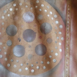 Puede incluir: Una chaqueta de cuero marr&oacute;n con un dise&ntilde;o de pintura en aerosol blanco y gris. El dise&ntilde;o presenta un c&iacute;rculo con c&iacute;rculos m&aacute;s peque&ntilde;os en el interior y un patr&oacute;n de puntos alrededor del borde.