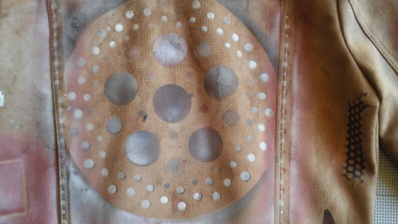 Puede incluir: Una chaqueta de cuero marr&oacute;n con un dise&ntilde;o de pintura en aerosol blanco y gris. El dise&ntilde;o presenta un c&iacute;rculo con c&iacute;rculos m&aacute;s peque&ntilde;os en el interior y un patr&oacute;n de puntos alrededor del borde.