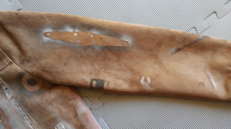 Puede incluir: Una chaqueta de ante marr&oacute;n con varios dise&ntilde;os de salpicaduras de pintura plateada y blanca. La chaqueta tiene un parche en la manga con una placa de metal.