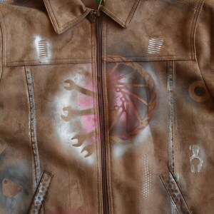 Puede incluir: Chaqueta de gamuza marr&oacute;n con dise&ntilde;os de pintura en aerosol blanca de herramientas y un engranaje. La chaqueta tiene una cremallera y un cuello.