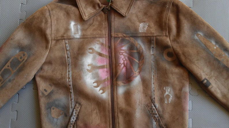 Puede incluir: Chaqueta de gamuza marr&oacute;n con dise&ntilde;os de pintura en aerosol blanca de herramientas y un engranaje. La chaqueta tiene una cremallera y un cuello.