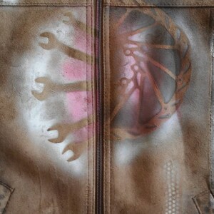 Puede incluir: Una chaqueta de ante marr&oacute;n con cierre de cremallera y un dise&ntilde;o pintado con aerosol en la parte delantera. El dise&ntilde;o presenta un patr&oacute;n abstracto rosa y blanco en el lado izquierdo y un patr&oacute;n abstracto marr&oacute;n y blanco en el lado derecho.